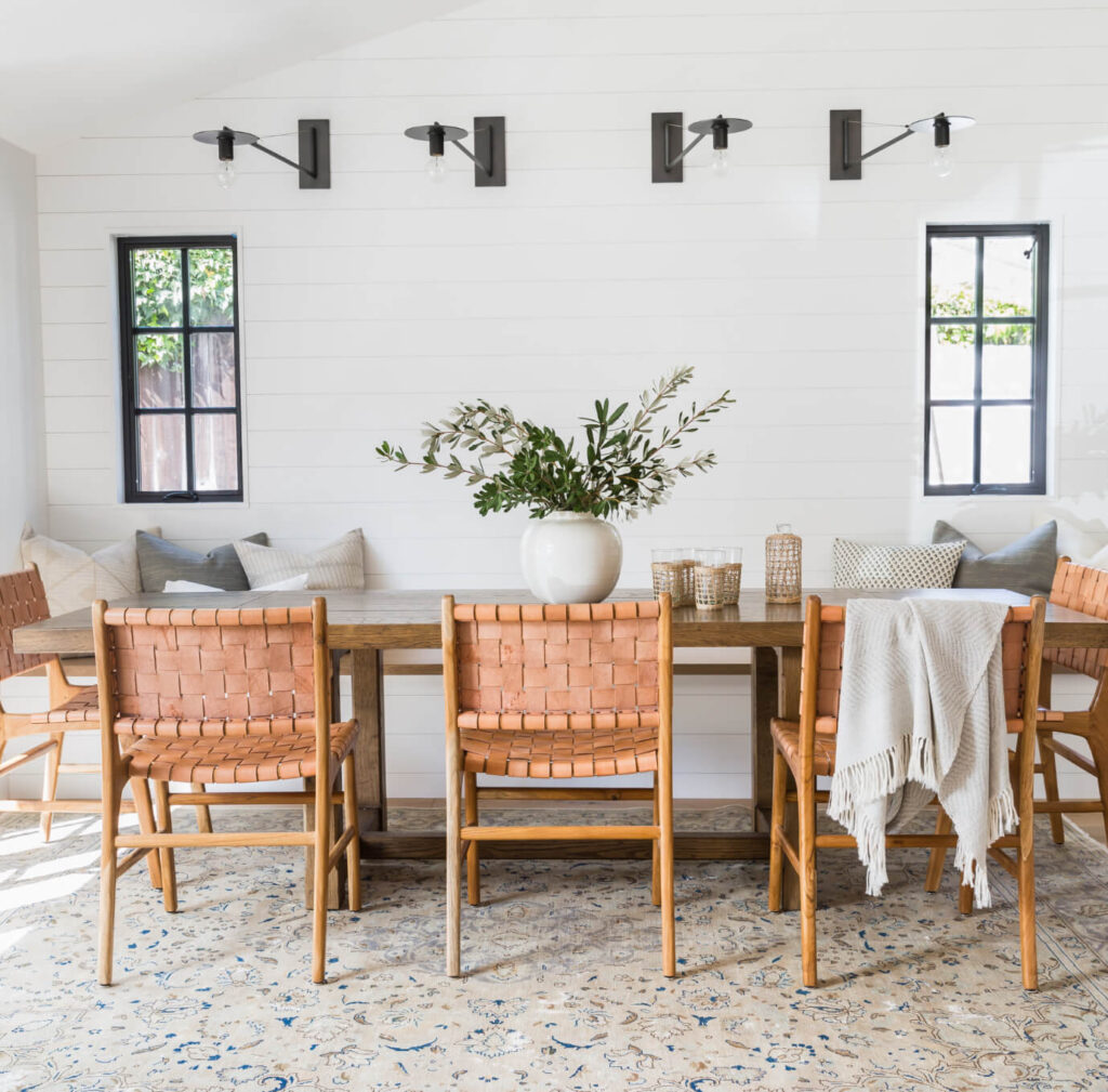 Modern farmhouse dining room with Scandinavian woven leather dining chairs around a wooden table on a vintage rug, shiplap walls and modern black metal farmhouse sconces